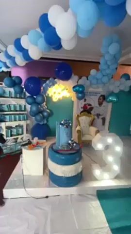 Here's a description of the video:

The video showcases a decorated party space, likely for a child's birthday. The predominant color scheme is blue and white, featuring balloon arches. A tiered cake decorated in blue tones is displayed on a platform. A small ornate chair sits in front of a poster featuring a child in a racing suit. A large illuminated "3" indicates the age. The room includes clear bubble chairs and an "Arsenal" sign in the background. Multiple children are present, dressed in party attire. The general atmosphere indicates a celebratory event.