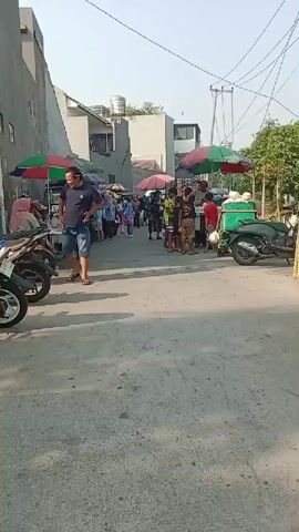 The video shows a narrow street lined with market stalls under colorful umbrellas. Several people are browsing the stalls or walking through the street. Some motorbikes are parked along the side of the street. In the foreground, a person on a motorbike is riding away from the camera. The scene appears to be a busy outdoor market on a sunny day.