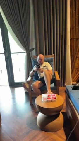 A man with a bald head, wearing a white t-shirt and black shorts, sits barefoot in a wooden armchair with a blue cushion. He holds a black smartphone and gestures with his left hand. A small round wooden table with a white box of red items is in front of him. The room has dark wooden floors and light brown curtains covering large windows.