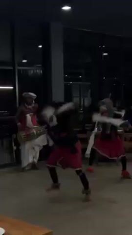 A video shows a group of four performers in a large, dimly-lit room with modern décor, which includes large windows. Two of the performers are drummers, dressed in traditional white garb and hats with red accents. They play small drums that are slung around their necks. The other two performers, presumably dancers, are dressed in black tops and red skirts. They dance, moving in a frenetic manner and spinning. One of the dancers wears a mask and has long white cloths in their hands. The dance is performed on a beige tile floor, and there are tables and chairs in the background. The scene appears to be a cultural performance at a restaurant or event.