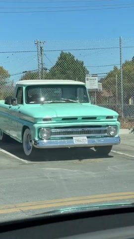 A light blue pickup truck with a white roof is parked in a parking lot. The truck has whitewall tires and a chrome bumper. A license plate with the number 7986223 is visible on the front of the truck. The truck is facing forward and appears to be in good condition. The background shows a chain-link fence, some trees, and a clear blue sky. The video is very short and the truck is stationary.