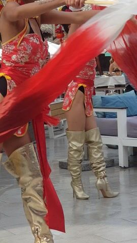 Two women stand side-by-side, facing the camera. They wear matching red dresses with floral patterns, gold trim, and red ties. Each dress is quite short, revealing black undergarments. They also wear tall, shiny gold boots. They are both holding open folding fans that are red and white. The women wear elaborate makeup and have their hair up, adorned with red and gold decorations. The background appears to be an outdoor restaurant or bar setting, with tables, chairs, and bamboo accents visible. Other people can be seen in the background. The camera angle is rotated so the image appears vertical, and the camera is close to the subjects.