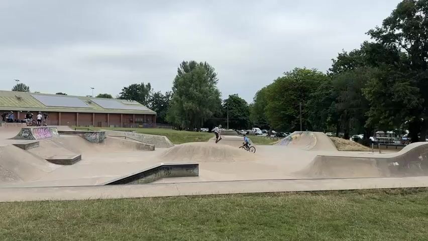 The video shows a skate park on an overcast day. In the background is a long, low building with a sloped roof, possibly a community center, with solar panels visible on the roof. There are trees behind the building and to the right of the frame. The skate park itself is made of concrete and features various ramps, bowls, and ledges. In the foreground, on the left, a group of people, some with bicycles, are standing near a set of ramps. In the middle ground, two individuals are actively using the skate park. One person is on a bicycle, riding on a curved ramp. Another person is on a scooter, also on a ramp. The ground around the skate park is covered in grass. The overall impression is a recreational space being utilized for sports.