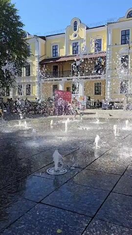 A sunny day with clear blue skies, a group of people are enjoying a fountain in a plaza. Water sprays from multiple spouts, creating a refreshing atmosphere. In the background, a yellow building with decorative trim and several windows stands prominently. A Ukrainian flag is visible on the building. Several people are walking around and interacting with the fountain, while others stand by. The ground is paved with dark, reflective tiles. Some trees are also visible, adding to the outdoor scene.