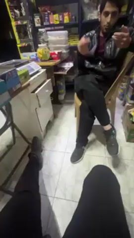 The video shows two individuals in what appears to be a small shop or store. One person is seated on a chair, wearing a black leather jacket, black pants, and black sneakers. He is gesturing with his hand as he talks. The other person's legs are visible in the foreground, wearing black pants and grey sneakers. The shop is stocked with various items on shelves and on a table. The floor is covered in white and grey patterned tiles. A white desk with a partially open drawer is also visible.