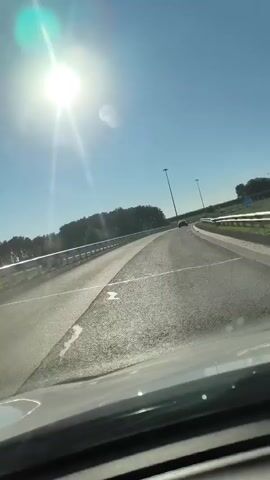 A first-person perspective video shows a bright sunny day from the perspective of a car driving on a highway. The sun is high in the clear blue sky, casting lens flares across the windshield. A metal guardrail runs along the left side of the road, and beyond it, trees form a dark green line against the horizon. In the distance, two tall lamp posts are visible on the right side of the highway. The road itself is asphalt with visible white lane markings. A single car can be seen driving in the distance on the right side of the highway. The car is moving forward at a steady pace.