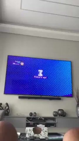 A person is viewing a television screen displaying a word-finding game. The game interface shows "Time's up!" and then changes to show the player found 26 words. The game displays a grid of letters: OAQE, RBTG, LIBA, AMCB. The player's avatar, labeled "Salmah," is in the lower left corner. Below the TV is a gray console cabinet with various decorative objects, including figurines, small globes, and a vase. The ceiling above is white with a recessed panel. Only the person's knees are visible in the foreground.