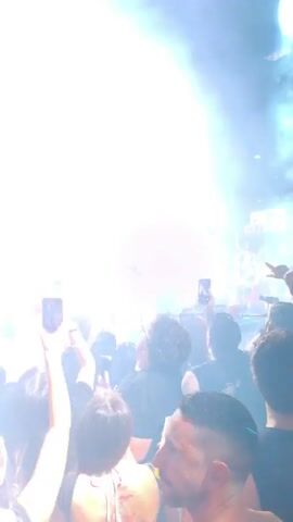 A concert with a large crowd. The stage is lit with bright lights and smoke. People in the crowd are holding up their phones to record the event. The performers on stage are silhouetted against the lights.