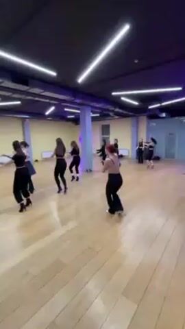 A group of about 10 women are dancing in a studio. They are all dressed in black, and the studio has a wooden floor. The room is well-lit with fluorescent lights, and there is a large pillar in the room. The women are performing a choreographed dance.