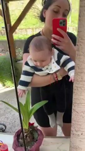 A woman with dark hair pulled back in a bun holds a baby in her arms as she takes a selfie in a mirror. The woman is wearing a black t-shirt and black shorts. The baby is wearing a striped blue and white sweater, black pants, and grey socks. The woman is holding a red smartphone. In the foreground to the left of the frame is a potted plant with green leaves. Behind the woman, visible through the mirror, is a partially obscured outdoor area with grass and a concrete surface. The camera is stable and focuses on the reflection in the mirror. The baby makes sounds.