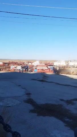 The video shows a concrete area with a view of a shipyard in the distance on a clear, sunny day.  A set of power lines cross the upper part of the frame. To the left, a chain fence runs along the edge of the frame. In the distance, there are several ships, mostly red and white, seemingly in dry dock. Two people are seen sitting on a structure towards the center of the frame. The concrete area in the foreground is cracked and has some patches of dark, damp spots. The landscape behind the shipyard appears flat and expansive.