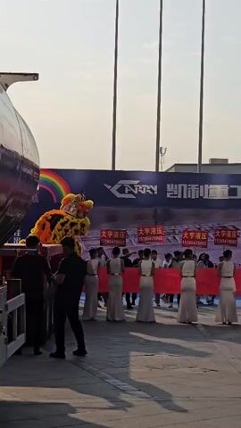 A group of people are standing in front of a large screen with the word CARRY on it. There are several women in long white dresses holding a red banner. Two lion dancers, one in yellow and one in red, are performing. A truck is also visible on the right side of the frame. The sky is clear and the sun is shining.