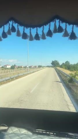 The video shows a POV from inside a vehicle, looking out the windshield. The view is of a two-lane road stretching into the distance under a clear blue sky. The road is bordered by a guardrail on either side, and beyond that, dry grass and some trees. In the distance, there are some houses and buildings. Hanging from the top of the windshield are decorative tassels and a Scania logo. Two black dice with white logos are hanging from the ceiling of the vehicle, in front of the windshield. The sun appears to be shining, casting shadows. The video appears to be a short clip of travel.