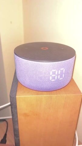A close-up shot shows a round, purple smart speaker with a digital display resting on top of a wooden speaker cabinet. The smart speaker has a black top and a textured, fabric-covered body. The digital display on the speaker shows a blurred pattern, indicating that the time or other information is changing rapidly. The wooden speaker cabinet beneath is a light brown color with visible wood grain. A black speaker with a gray front is partially visible to the left of the wooden cabinet. Electrical cords are visible on the floor, suggesting the setup is functional. The background consists of an off-white wall. The camera slightly moves around the smart speaker, showing different angles of it. Music is playing in the background.