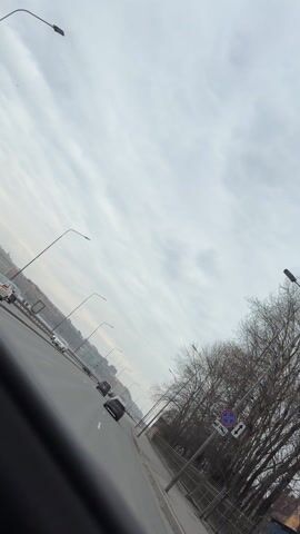 The video opens with a slanted view of a bridge highway, showing multiple lanes filled with cars. The sky is overcast with textured grey clouds. Lampposts line the highway. Trees with bare branches stand to the right. The perspective shifts inside a car, revealing the driver, a fair-skinned man with short hair. The car is a convertible with black interior and a white exterior. The driver is holding the steering wheel. A small, decorative skeleton hangs from the rearview mirror. A navigation system is visible on the dashboard screen. The camera focuses on the driver and the interior of the car as it drives on the highway.