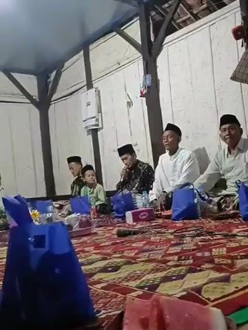 Four individuals, appearing to be males of varying ages, are seated on the floor, which is covered by a red and white patterned rug. The individuals are arranged from left to right in the frame. The person second from the left is a young boy wearing a light green long-sleeved shirt and a black skullcap. To his right, a man in a black and white patterned garment and a black skullcap is holding a microphone and speaking. To the far right, an older man in a white shirt and a black skullcap is seated. To the left of the young boy is another young male wearing a black and white patterned garment and a black skullcap. In front of the seated individuals are several blue bags, water bottles, and a tissue box. The background consists of a light-colored wooden wall.