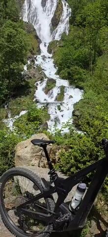 The scene shows a black mountain bike parked on a rocky surface with lush greenery in the foreground. Behind it, a large, cascading waterfall flows down a rocky slope, surrounded by trees.  At least two people are visible on the rocks within the waterfall. The camera pans up to better capture the waterfall. The setting appears to be a scenic, mountainous area.