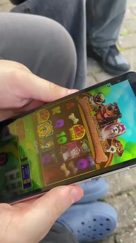 A person wearing grey pants and blue Crocs is playing a dog-themed slot machine game on their phone. Another person wearing dark jeans and black shoes is seated next to them. The game on the phone features dog characters, bones, and gem symbols on a green background. The phone is positioned horizontally in the person's hands as they play the game. The scene appears to be outdoors, with what looks like pavement visible beneath the person's feet.
