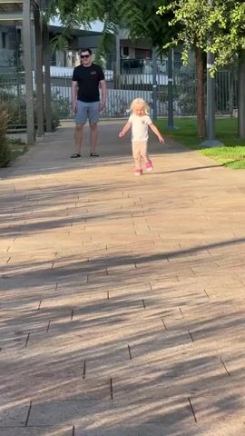 The video shows a young blonde child with fair skin running towards the camera on a paved walkway. The child is wearing a white t-shirt with a brown embroidered bear on the left chest, light pink leggings, and pink shoes. The child's blonde hair is partially visible. Behind the child, further down the walkway, a man wearing a black t-shirt, grey shorts, and sunglasses is walking away from the camera. He is wearing black flip-flops. The setting appears to be a park or a well-maintained outdoor area with trees and grass visible to the right of the walkway. There is also a fence and buildings in the background. The sun is casting long shadows from the trees onto the walkway, suggesting it is late afternoon or early evening. The overall mood seems to be pleasant and relaxed, with the child appearing energetic and playful. There is no visible text in the video.