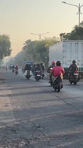 A group of motorcyclists are riding in formation on a paved road. The motorcyclists are all wearing helmets, and some are wearing backpacks. The road is lined with trees, and there is a large white truck in the background. The sky is clear and the sun is shining. The video appears to have been filmed during the daytime.