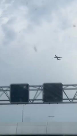 The video shows a blurry view of a highway overhead sign structure against a cloudy sky. The sign structure is made of metal beams and has two large black rectangular signs attached to it. Below the sign structure is a white concrete barrier. A street light is visible on the left side of the frame. The camera appears to be moving, as evidenced by the slight blur in the image. The overall environment appears to be a typical highway scene.