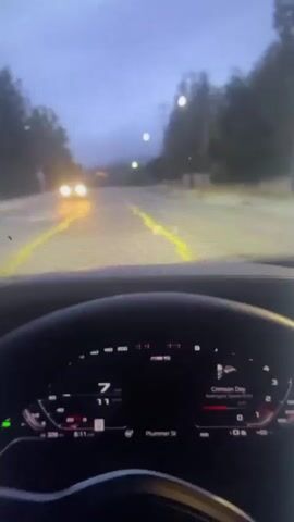 The video shows a first-person view from inside a car. The driver's digital dashboard is visible, displaying speed, gear selection, and navigation information. The car is on a road at dusk or night, with headlights of an oncoming vehicle visible in the distance. The road has yellow lane markings. Trees and foliage line the sides of the road. The sky is dark and overcast. The car appears to be moving.