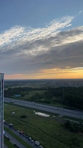 An aerial, upward-looking video captures a wide shot of a suburban landscape at dusk. The sky is a mixture of blue and gray clouds, with the sun setting on the horizon, casting a warm, golden glow. Below the sky, a dense green forest stretches across the middle ground, with a highway running through it. Cars can be seen driving on the highway. In the foreground, to the left, is a portion of a tall building, and to the right, a grassy area with a few parked cars. The overall impression is one of a peaceful evening in a developing urban area.