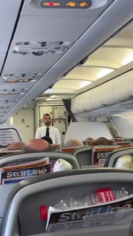 This video shows the interior of an airplane cabin. In the foreground, the backs of airplane seats are visible, with magazines and a plastic bottle with a red cap in the seat pockets. In the mid-ground, the heads of passengers are visible, and further back, a male flight attendant in a white shirt and dark tie stands near a doorway that leads to the galley area. The ceiling of the cabin features overhead compartments and lighting. The overall environment appears to be during a flight or before takeoff, as indicated by the presence of the flight attendant and the seatbelt signs illuminated above.
