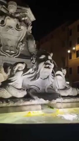 The video shows a detailed view of a fountain in Rome at night, specifically focusing on the sculptural elements that decorate it. The fountain is brightly lit, highlighting the elaborate carvings that include a central human-like face with exaggerated features, possibly a grotesque or a mythological figure. Water is flowing from the mouth of this figure into the pool below. The sculpture is made of white stone, likely marble, and is part of a larger column or monument. Behind the fountain, a building is visible, featuring multiple windows and lit by street lamps, suggesting an urban setting. The scene is captured at a close angle, emphasizing the intricate detail of the fountain and its surroundings.