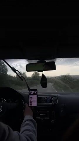 The video is taken from the driver's seat of a car, looking out through the windshield. Rain is falling, and the wipers are in motion. The driver's hands are visible on the steering wheel, and a smartphone is mounted in the center console, displaying a music playlist. A black car passes in the opposite direction. The environment outside the car appears to be a rural road surrounded by trees. The scene is somewhat dark due to the weather and time of day.