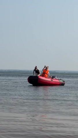 A red inflatable boat with a black stripe is on the water. There are three people in and around the boat. One person is lying face down on the front of the boat. Two people are sitting in the boat. One person in the boat is wearing an orange life jacket. Another person in the boat is wearing a white hat. A third person is partially submerged in the water to the left of the boat. The sky is clear and blue. The water is calm.