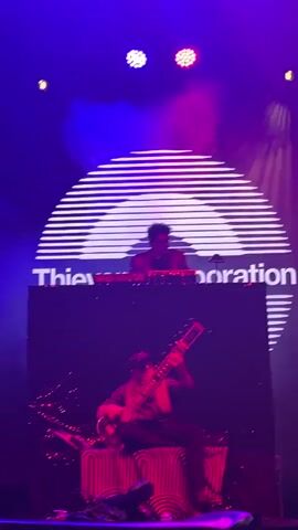A live music performance is captured in this video, featuring two performers on a dimly lit stage. The stage is set with a large circular backdrop displaying a white, stylized sun or radar. Below the backdrop, a black platform with a digital screen displays a pattern of red, glowing stripes. On the platform, a performer stands behind a DJ console, wearing glasses and a jacket, operating electronic music equipment. In front of the DJ, a second performer is seated, playing a sitar, an Indian string instrument. The performer on the sitar is wearing a cap and sunglasses. The stage is illuminated by vibrant purple and pink stage lights, with some red and blue lights also visible. Smoke or fog adds to the atmospheric effect. Towards the end of the video, a third person, a woman with purple hair, appears to be dancing on the left side of the stage. The audience is visible at the bottom of the frame, with some people holding up their phones to record the performance.