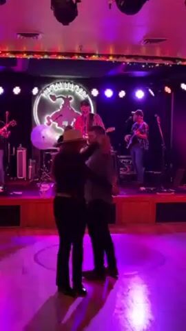 A video captures a band performing live at "The Cowboy Bar," with a couple dancing in the foreground. The band consists of four members on a raised stage, with the band's logo displayed behind them. Stage lighting illuminates the band members and the couple. The couple is seen dancing in front of the stage. The woman is wearing a cowboy hat, a dark jacket, and dark pants, while the man is in a dark shirt and dark pants. The dance floor is marked with a circular design. The scene is lit with colored lights, mostly purple and pink. A tip jar is visible near the edge of the stage.