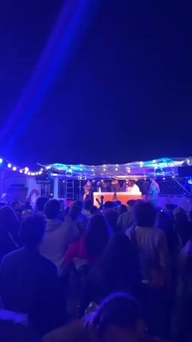 A short video depicts a lively party at night. A band of about six members performs on a stage adorned with string lights. The stage appears to be set up on a vehicle, perhaps a trailer or bus. A camera is positioned in front of the performers, potentially recording the event. The crowd, consisting of numerous individuals, faces the stage, some with their arms raised, appearing to enjoy the music and ambiance. The atmosphere is illuminated by blue stage lights, enhancing the night's party atmosphere.