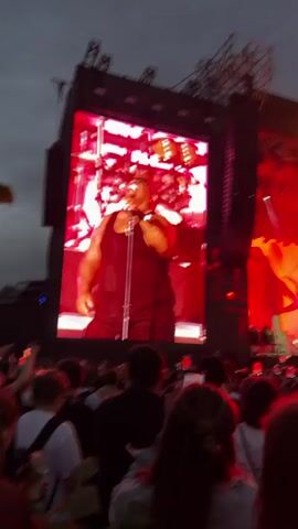 The video shows a wide shot of a large outdoor concert crowd viewed from behind. The crowd is facing a large stage with a massive LED screen dominating the left side of the frame. On the screen, a bald man with tattoos, wearing a black tank top and sunglasses, is singing into a microphone. Behind him, on the actual stage, a drummer can be seen. The stage is lit with vibrant blue and red lights, and the words "PARK LIVE" are displayed in large illuminated letters above the stage. People in the crowd are holding up their phones, presumably to record the performance. The sky appears overcast.