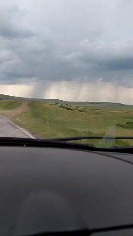 The camera is positioned inside a vehicle, looking forward through the windshield. The top of the dashboard and the bottom of the windshield are visible in the foreground. A two-lane road stretches ahead, appearing wet and transitioning from pavement to dirt or gravel. Green, undulating fields extend on both sides of the road. In the distance, a triangular road sign is visible on the right side of the road. The sky is overcast with dark grey clouds, and distinct streaks of rain are falling from the clouds over the distant landscape. The sound of the vehicle moving, possibly over a wet or unpaved surface, is audible.