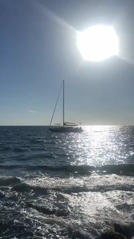 A sailboat is on the water under a bright sun. The sun is in the upper right corner of the frame, causing a glare on the water. The water is choppy and the waves are breaking on rocks in the foreground. The sky is clear and blue.