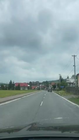The video shows a first-person perspective driving on a paved road through a small town on an overcast day. The road has two lanes separated by a solid white line. There are houses and buildings on either side of the road, most with red roofs. Utility poles and wires run along the side of the road. There is a pedestrian crossing marked by white stripes on the road. A dark colored car is visible driving in the distance. The sky is filled with heavy gray clouds.