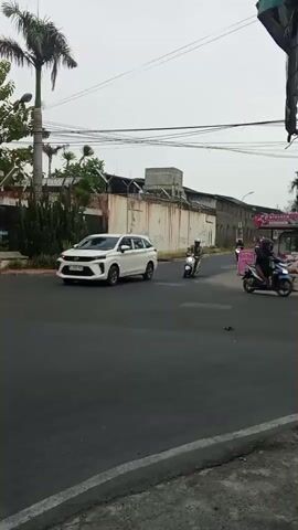 A white SUV is seen driving down a road. Several motorcycles are also on the road, some with riders, some without. There are buildings and trees in the background. The sky is overcast.