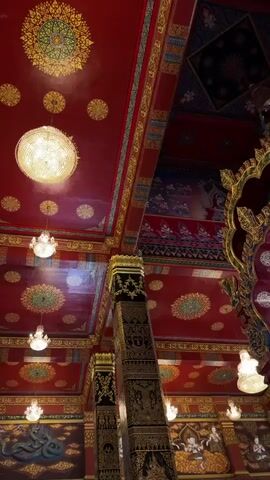 This is a close-up, low-angle, shaky, vertical video of the interior of a temple. The ceiling is a deep red color and is elaborately decorated with gold patterns and circular motifs. Several chandeliers and pendant lights hang from the ceiling, emitting a bright, warm light. Ornate pillars, also decorated with gold patterns, rise up into the frame. Murals depicting figures and scenes are visible on the walls below the ceiling. The overall impression is one of opulence and grandeur.