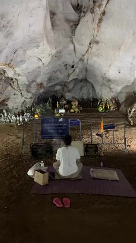 A woman is meditating inside a cave. The woman is sitting cross-legged on a purple mat, facing an elevated area decorated with various statues and shrines.  She is wearing a white t-shirt and tan pants. Pink flip-flops sit beside the mat. A bottle of water and a brown shopping bag are near the mat. Several signs with Thai writing are positioned between the woman and the elevated area. The cave has a high ceiling with a rough, textured surface. The ground appears to be a dark, earthy color.