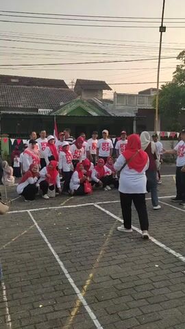 A group of people, most wearing white shirts with red lettering that says "RT 05" and red headscarves or hats, are gathered in an outdoor paved area marked with white lines. Some individuals are standing, while others are crouched or sitting on the ground. A woman in a white shirt and red headscarf is facing away from the camera, and another person in a similar outfit is to her right. In the background, there are houses and trees. The overall impression is of a community event or gathering.