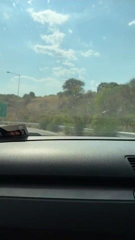 The video shows a view from inside a car driving on a sunny day. The dashboard is in the foreground. Through the windshield, a highway is visible with a guardrail on the right. A street lamp and a green highway sign are also visible to the left. The landscape consists of hills covered with dry grass and scattered trees. The sky is blue with scattered white clouds. There are some spots on the windshield.