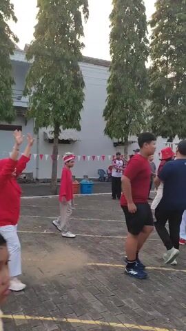 A group of people are gathered outdoors on a paved surface marked with white lines. The scene appears to be a street or courtyard with buildings and trees in the background. Several individuals are wearing red shirts and white pants, some with red headbands or hats. One person in the foreground, wearing a red shirt with "INDONESIA" written on it, is speaking to another person wearing a black t-shirt. In the background, a flag that appears to be the Indonesian flag is visible. The lighting suggests it is either early morning or late afternoon. The overall atmosphere seems festive or celebratory, possibly related to a local event or competition.