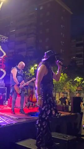 A live band is performing on an outdoor stage at night. A male singer, wearing a black cowboy hat, a black vest, and black pants adorned with skull patterns, is holding a microphone and singing. To his left, a bald male guitarist, wearing a black vest and jeans, is playing an electric guitar. Several other guitars and music equipment are visible on the stage. In the background, buildings are lit, and trees are present. The stage is illuminated by purple and red lights. Some audience members are visible in the lower right corner of the frame.