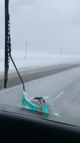 The view from inside a vehicle driving on a rain-swept road on a grey day. A partially crushed package of chewing gum sits on the dashboard. Raindrops cover the windshield, and the wipers are moving. Power lines run alongside the road. Visibility is poor due to the rain and fog.