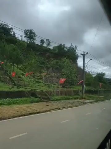 A motorbike rides along a paved road bordered by a grassy area with red flags. Behind the grassy area, a steep, vegetated hillside rises, with some trees and power lines visible. The sky is overcast.
