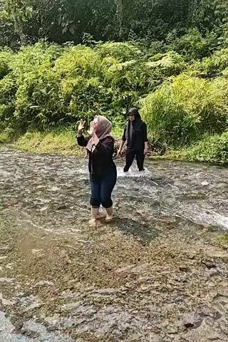 Two women are walking in a shallow river. The woman in the foreground is wearing a light-colored hijab, a black shirt, and dark pants. The woman in the background is wearing a black hijab and a black outfit. They are surrounded by lush green vegetation on either side of the river. The riverbed is rocky and the water is clear. The sun is shining, indicating it is daytime.