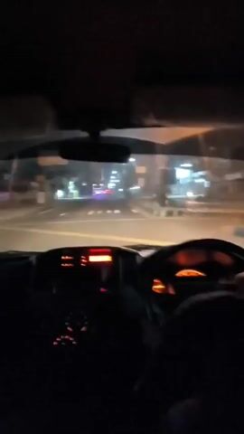 The video shows a first-person view from the inside of a vehicle driving at night. The dashboard is visible with illuminated orange and red lights. The road ahead is lit by headlights and streetlights. There are other vehicles on the road, including a truck with flashing blue and red lights in the distance. Buildings and trees line the side of the road. The camera perspective is static, looking forward through the windshield.