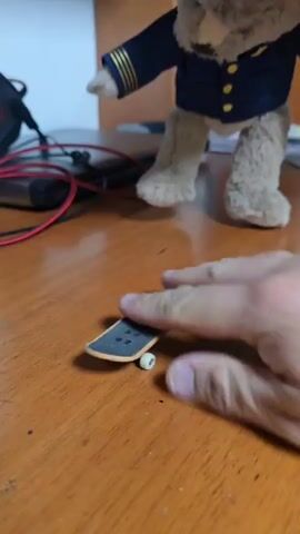 A video shows a hand placing a fingerboard on a wooden table. Behind the fingerboard, a plush toy bear is visible wearing a navy blue pilot uniform. The hand attempts to manipulate the fingerboard to do a trick, but it mostly stays in place. The bear sits still in the background. A red cable and some small black devices are also present on the table.